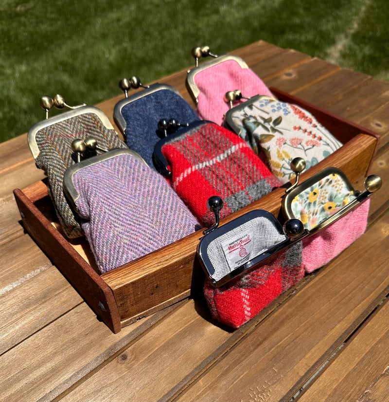 A collection of Aberdeen Kiss Lock Pouches in various Harris Tweed patterns and colors, with a solid leather finish, displayed in a wooden tray.