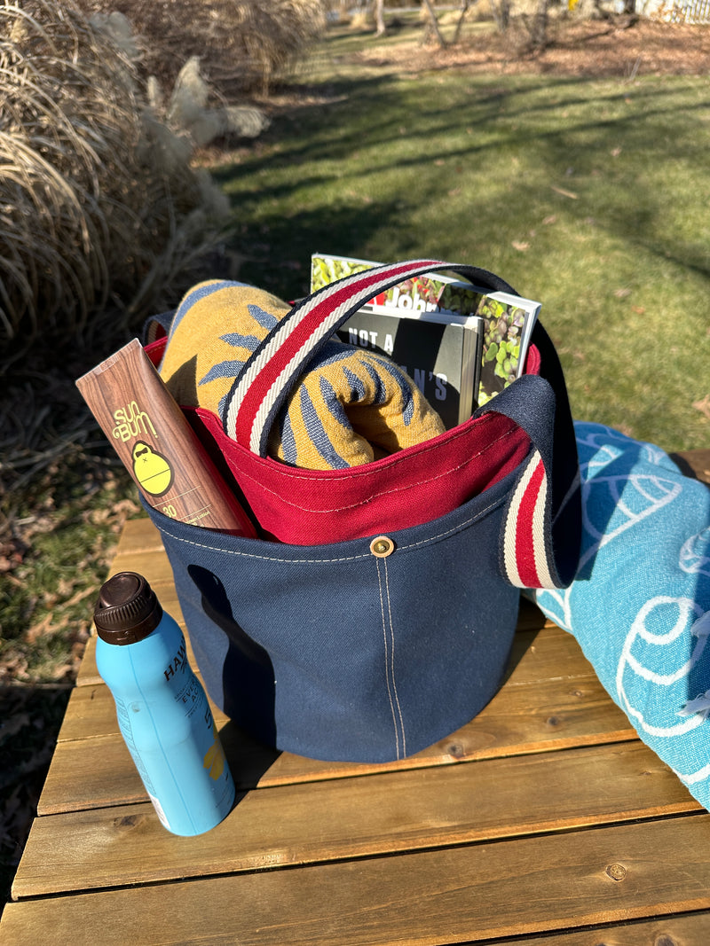 Close up of the Waterman Bucket Shorty, a small navy blue and red canvas bucket tote with long webbing shoulder straps with beach towel, with magazine and sun tan lotion. A versatile beach or pool bag handcrafted from 18oz cotton duck canvas for a classic nautical aesthetic