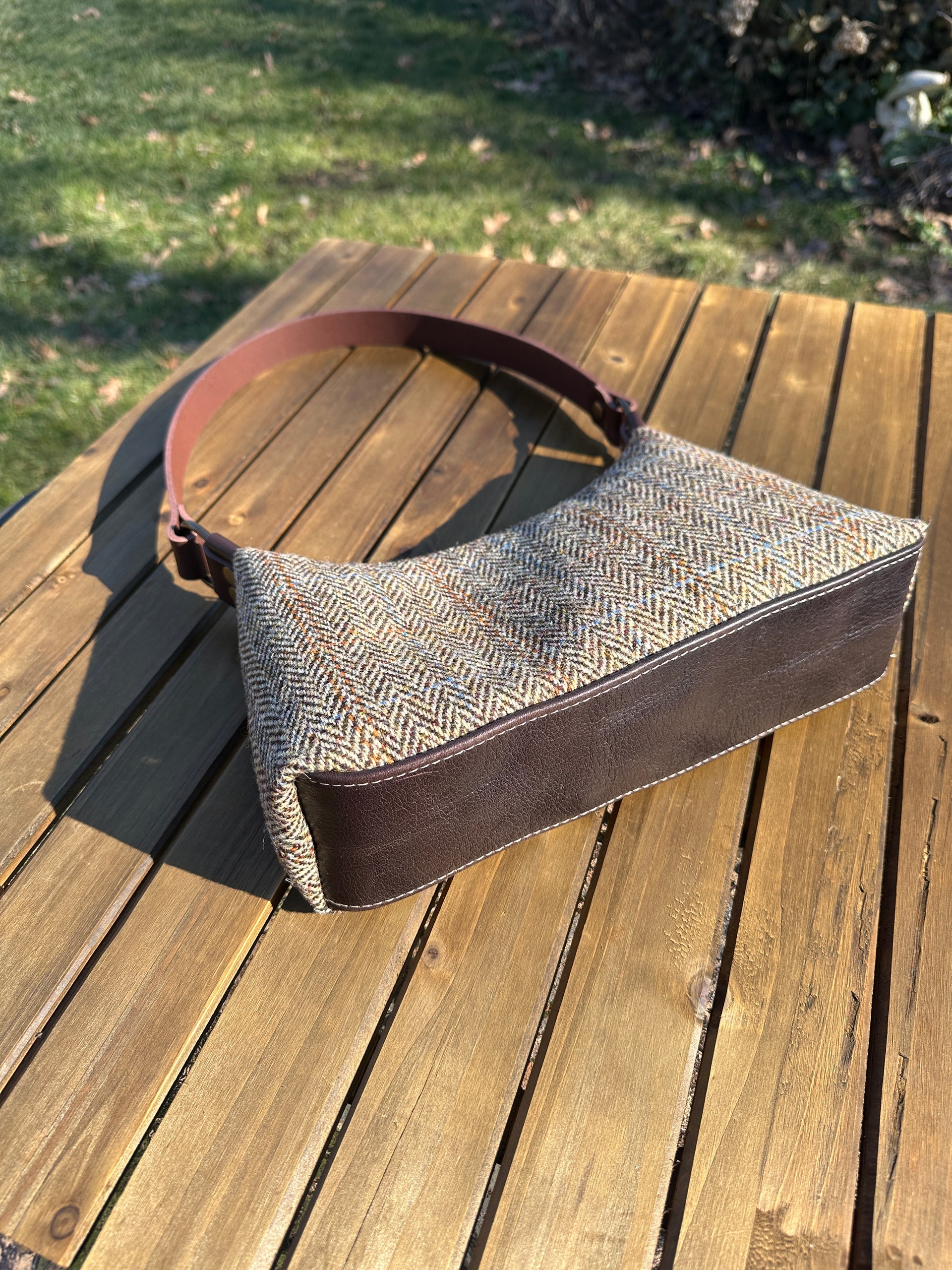 Close-up of the bottom of 'The Isla' bag showing the structured dark brown leather base with white contrast stitching. The bag is shown from a low angle on a wooden slatted table, highlighting the depth and the Harris Tweed fabric.