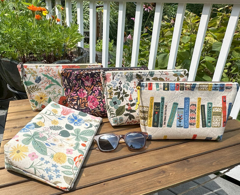 A collection of large Canvas Schooner Pouches in various Rifle Paper Co. Canvas patterns, displayed on a wooden table with sunglasses on one of them.