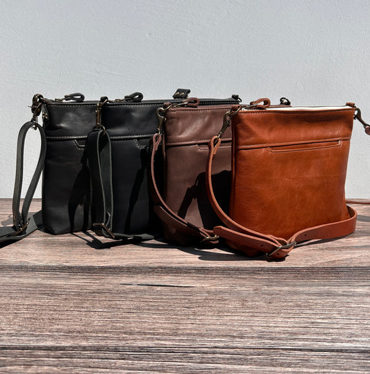 Group photo showcasing four handcrafted Leather Bayside Crossbody bags, each made with Horween leather in unique finishes. Bags are arranged to show variations in leather color and grain—Sienna, Vintage Dark Brown, Bomber Jacket Black, and Rustic Charcoal—each featuring solid brass hardware, a full metal zipper, and an adjustable vegetable-tanned leather strap. Designed and made in Maryland.