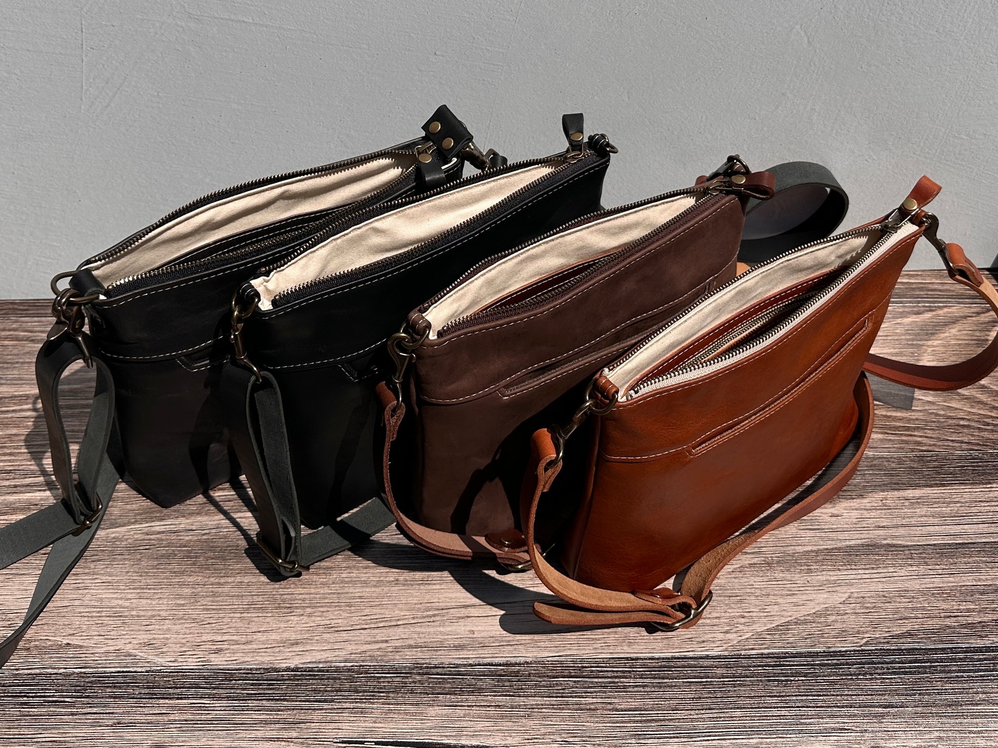 Overhead group shot of four open handcrafted Leather Bayside Crossbody bags, showcasing their unique Horween leather finishes: Sienna, Vintage Dark Brown, Bomber Jacket Black, and Rustic Charcoal. Each bag displays its open interior, solid brass hardware, full metal zipper, and adjustable vegetable-tanned leather strap. Designed and made in Maryland.