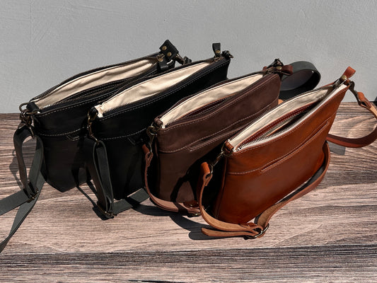 Overhead group shot of four open handcrafted Leather Bayside Crossbody bags, showcasing their unique Horween leather finishes: Sienna, Vintage Dark Brown, Bomber Jacket Black, and Rustic Charcoal. Each bag displays its open interior, solid brass hardware, full metal zipper, and adjustable vegetable-tanned leather strap. Designed and made in Maryland.