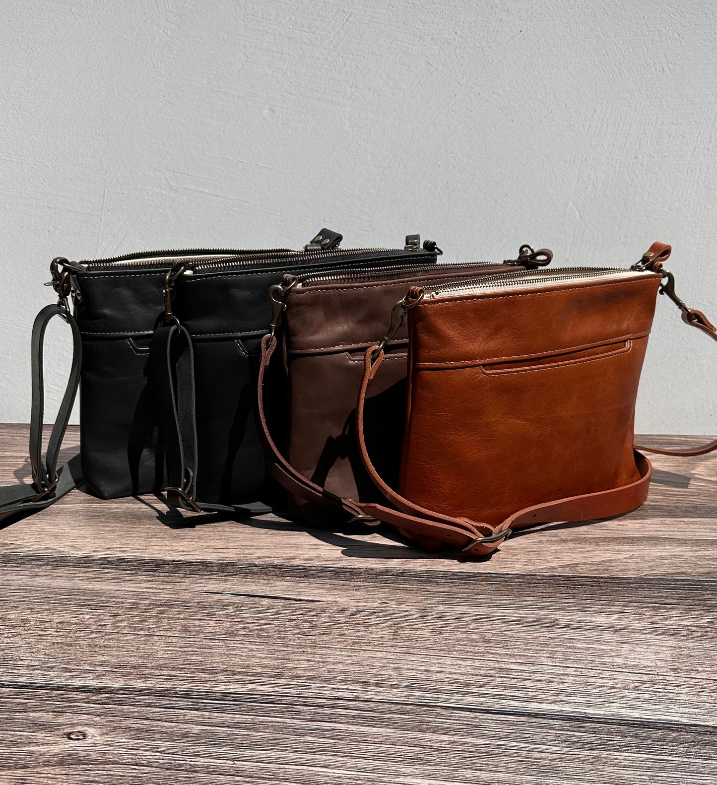Group photo showcasing four handcrafted Leather Bayside Crossbody bags, each made with Horween leather in unique finishes. Bags are arranged to show variations in leather color and grain—Sienna, Vintage Dark Brown, Bomber Jacket Black, and Rustic Charcoal—each featuring solid brass hardware, a full metal zipper, and an adjustable vegetable-tanned leather strap. Designed and made in Maryland.
