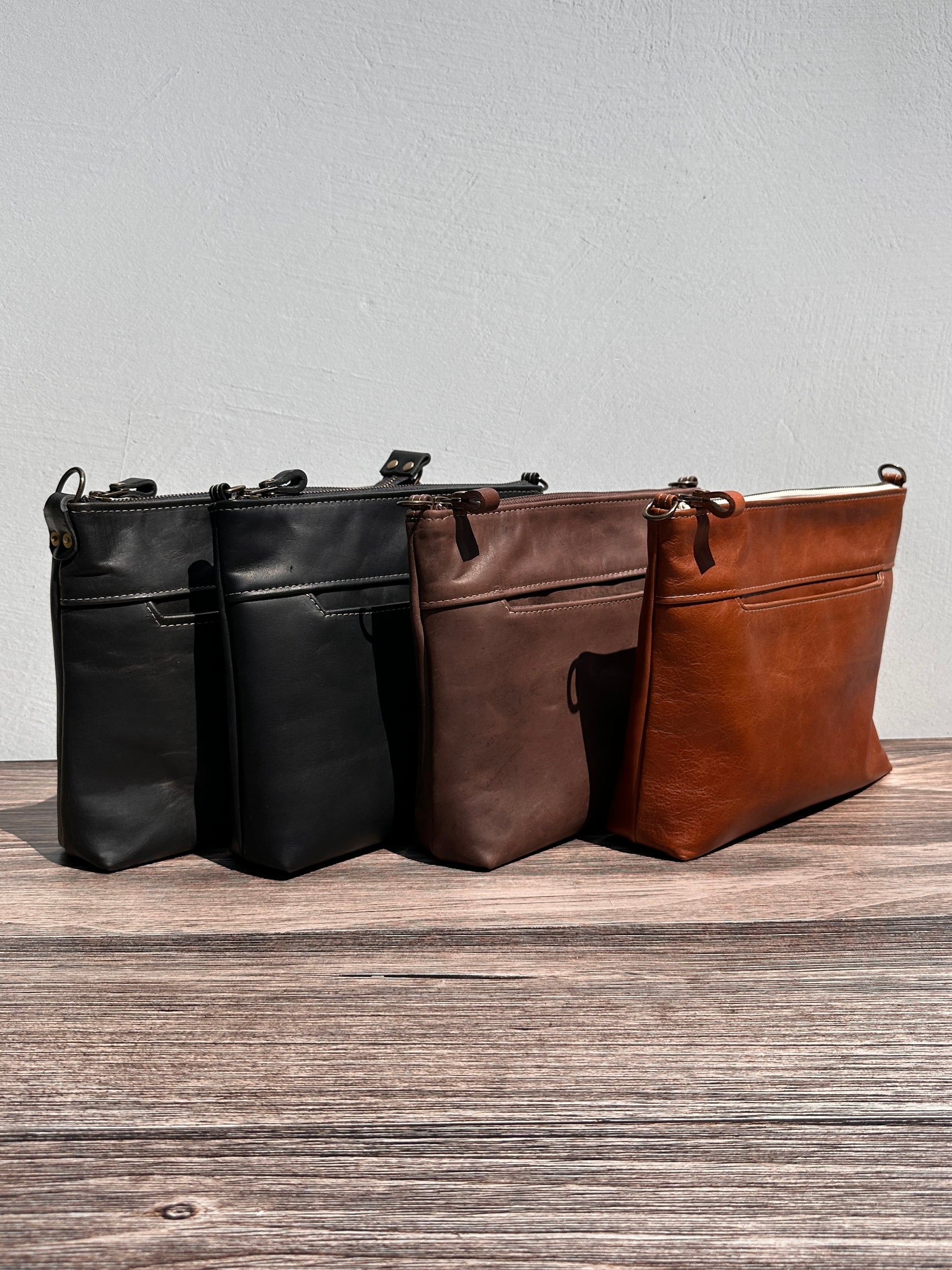 Group photo showcasing four handcrafted Leather Bayside Crossbody bags, each made with Horween leather in unique finishes. Bags are arranged to show variations in leather color and grain—Sienna, Vintage Dark Brown, Bomber Jacket Black, and Rustic Charcoal—each featuring solid brass hardware, a full metal zipper, and an adjustable vegetable-tanned leather strap. Designed and made in Maryland.