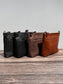 Group photo showcasing four handcrafted Leather Bayside Crossbody bags, each made with Horween leather in unique finishes. Bags are arranged to show variations in leather color and grain—Sienna, Vintage Dark Brown, Bomber Jacket Black, and Rustic Charcoal—each featuring solid brass hardware, a full metal zipper, and an adjustable vegetable-tanned leather strap. Designed and made in Maryland.