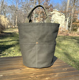 Large olive green heavy-duty canvas garden tool bag with four deep exterior pockets holding seed packets. Features durable webbing grab handles and a structured round base for upright utility.