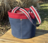 Waterman Bucket Shorty is a small navy blue and red canvas bucket tote with long webbing shoulder straps. A versatile beach or pool bag handcrafted from 18oz cotton duck canvas for a classic nautical aesthetic.