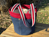Waterman Bucket Shorty is a small navy blue and red canvas bucket tote with long webbing shoulder straps. A versatile beach or pool bag handcrafted from 18oz cotton duck canvas for a classic nautical aesthetic.