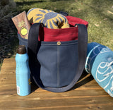 Close up of the Waterman Bucket Shorty, a small navy blue and red canvas bucket tote with long webbing shoulder straps with beach towel, with magazine and sun tan lotion. A versatile beach or pool bag handcrafted from 18oz cotton duck canvas for a classic nautical aesthetic