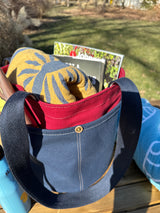 Close up of the Waterman Bucket Shorty, a small navy blue and red canvas bucket tote with long webbing shoulder straps with beach towel, with magazine and sun tan lotion. A versatile beach or pool bag handcrafted from 18oz cotton duck canvas for a classic nautical aesthetic