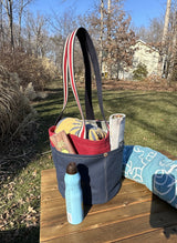 Close up of the Waterman Bucket Shorty, a small navy blue and red canvas bucket tote with long webbing shoulder straps with beach towel, with magazine and sun tan lotion. A versatile beach or pool bag handcrafted from 18oz cotton duck canvas for a classic nautical aesthetic