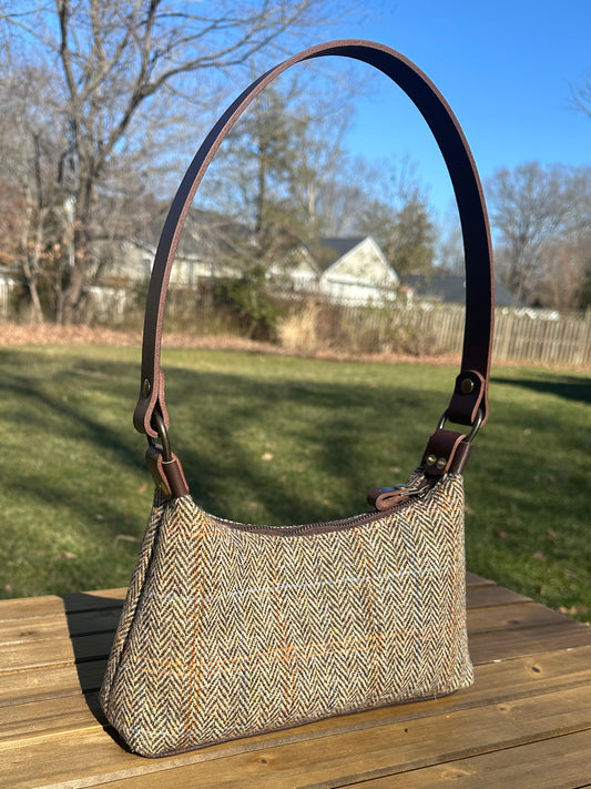 A handmade curved-top shoulder bag named 'The Isla' sitting on a wooden table outdoors. The bag features a brown herringbone Harris Tweed exterior with a slim, dark brown leather shoulder strap and brass hardware.