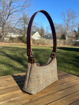 A handmade curved-top shoulder bag named 'The Isla' sitting on a wooden table outdoors. The bag features a brown herringbone Harris Tweed exterior with a slim, dark brown leather shoulder strap and brass hardware.