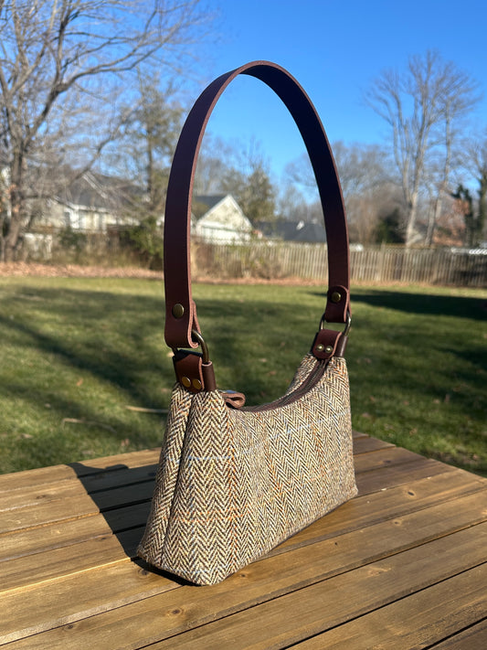 A handmade curved-top shoulder bag named 'The Isla' sitting on a wooden table outdoors. The bag features a brown herringbone Harris Tweed exterior with a slim, dark brown leather shoulder strap and brass hardware.
