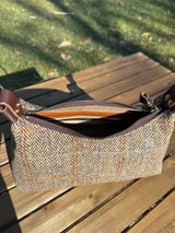 A handmade curved-top shoulder bag named 'The Isla' sitting on a wooden table outdoors. The bag features a brown herringbone Harris Tweed exterior with a slim, dark brown leather shoulder strap and brass hardware.