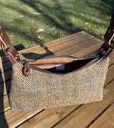A handmade curved-top shoulder bag named 'The Isla' sitting on a wooden table outdoors. The bag features a brown herringbone Harris Tweed exterior with a slim, dark brown leather shoulder strap and brass hardware.