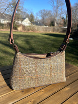 A handmade curved-top shoulder bag named 'The Isla' sitting on a wooden table outdoors. The bag features a brown herringbone Harris Tweed exterior with a slim, dark brown leather shoulder strap and brass hardware.