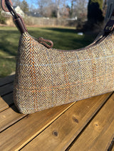 A handmade curved-top shoulder bag named 'The Isla' sitting on a wooden table outdoors. The bag features a brown herringbone Harris Tweed exterior with a slim, dark brown leather shoulder strap and brass hardware.