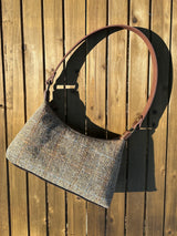 A handmade curved-top shoulder bag named 'The Isla' sitting on a wooden table outdoors. The bag features a brown herringbone Harris Tweed exterior with a slim, dark brown leather shoulder strap and brass hardware.