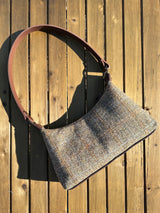 A handmade curved-top shoulder bag named 'The Isla' sitting on a wooden table outdoors. The bag features a brown herringbone Harris Tweed exterior with a slim, dark brown leather shoulder strap and brass hardware.