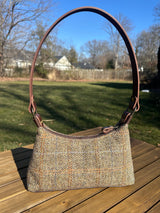 A handmade curved-top shoulder bag named 'The Isla' sitting on a wooden table outdoors. The bag features a brown herringbone Harris Tweed exterior with a slim, dark brown leather shoulder strap and brass hardware.